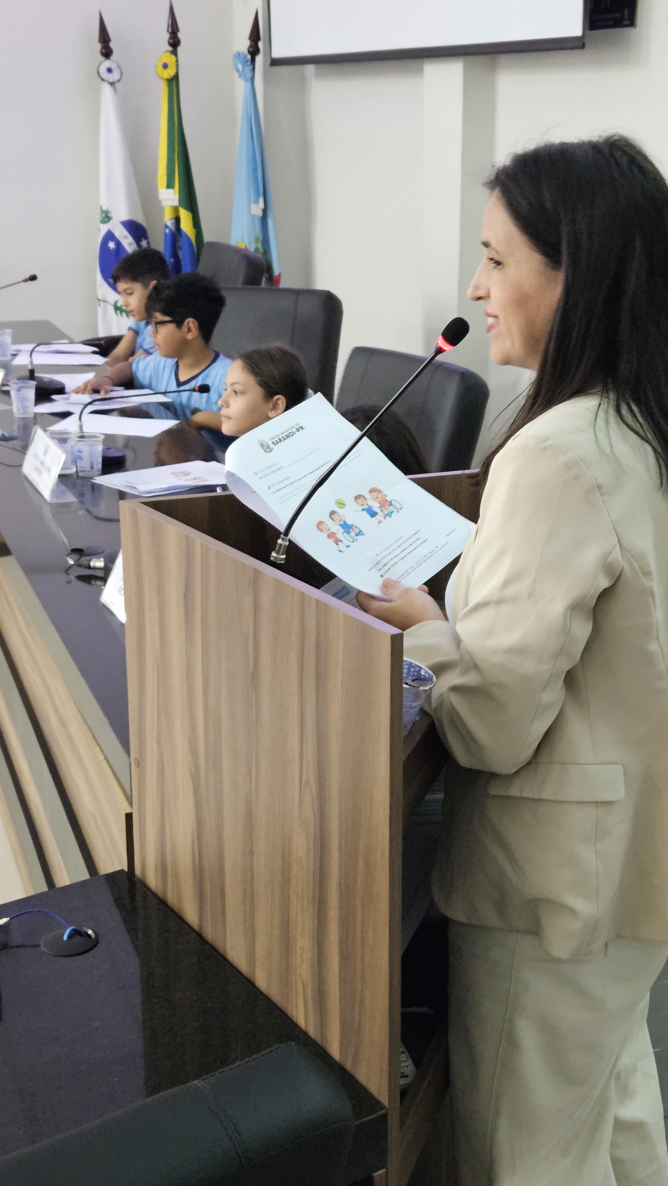 Professora fala ao microfone durante sessão da Câmara Mirim, enquanto Vereadores Mirins acompanham a atividade sentados à mesa. Ao fundo, estão as bandeiras oficiais.